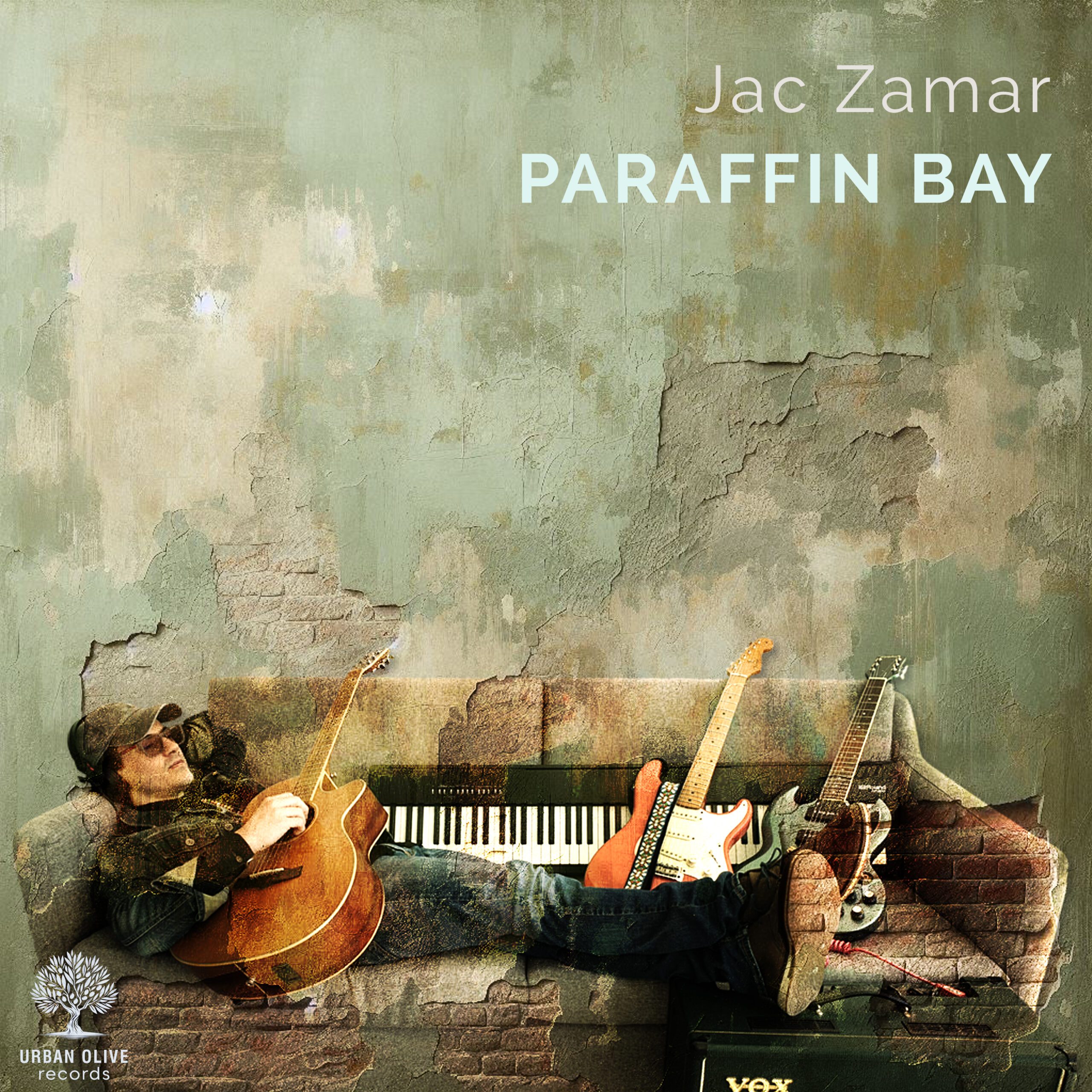 Paraffin Bay — debut album artwork: Jac Zamar reclining with multiple instruments on a worn couch against a textured painted wall
